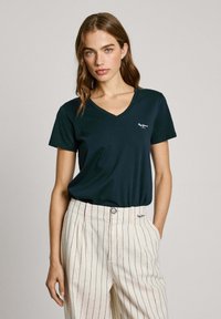 Mörkgrön V-ringad t-shirt i mjukt material, med en liten vit logotyp på vänster bröst, kombinerad med ljusrandiga, högmidjade byxor.