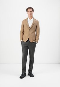 Blazer beige, pantalon texturé gris, chemise blanche et chaussures noires. Le blazer a deux poches sur le devant et une fermeture à un bouton.