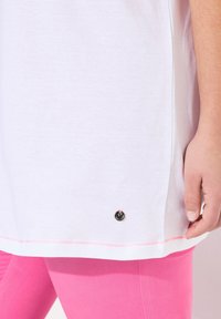 Weißes T-Shirt mit abgerundetem Saum, versehen mit einem schwarzen, runden Logo und einem pinken Akzent auf der unteren Seite, kombiniert mit pinken Leggings.