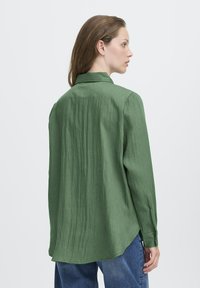 Femme de profil portant une chemise verte ample à manches longues avec un col et un jean bleu, sur fond blanc uni.