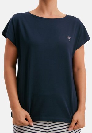 Laivastonsininen lyhythihainen paita, jossa on pieni valkoinen kukkaommellus vasemmassa rinnassa, puettuna raidallisten housujen kanssa, kädet pitävät paidan helmaa.