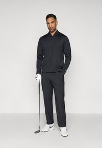 Άνδρας με μαύρη μακρυμάνικη μπλούζα και παντελόνι που κρατάει μία γκολφ κλαμπ, φορώντας λευκό γάντι γκολφ και λευκά αθλητικά παπούτσια με μαύρη λεπτομέρεια.