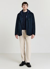 Marineblauer zweireihiger Mantel über einem marineblauen Pullover, kombiniert mit hellbeigen, weit geschnittenen Hosen und schwarzen Schuhen. Texturiertes Material und Kapuzen-Design.