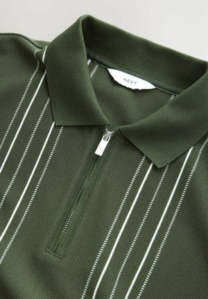 Polo shirt vert foncé avec une fermeture éclair sur le devant, mettant en avant des rayures verticales blanches et un tissu texturé. L'étiquette à l'intérieur indique "NEXT."