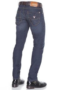 Donkerblauwe denim jeans met zichtbare stiksels, twee achterzakken met een geborduurde ontwerp, gedragen met een zwart leren riem en schoenen.