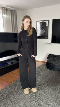 Chemise noire à boutons avec manches longues, associée à un pantalon noir à jambes larges. Chaussures de couleur claire complètent la tenue. Tapis gris doux et texturé.