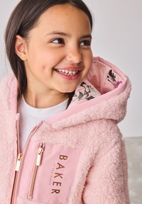 Ragazza sorridente con lunghi capelli scuri che indossa una giacca rosa soffice con cappuccio foderato a fiori e dettagli in oro sulle cerniere.