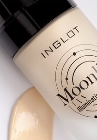 Beige flytande foundation i en klar glasflaska med ett svart pumplock. Etiketten har svart text och ett cirkulärt mönster. Utspilld produkt synlig.