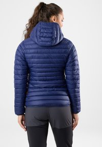 Marinblå pufferjacka med huva, med horisontella quiltade sektioner och en slät yta. Inkluderar en liten logotypdetalj vid fållkant.