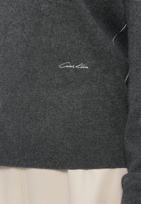 Ciemnoszary sweter z dzianiny z prążkowanymi mankietami i dołem, z białym logo "Calvin Klein" wyszytym z przodu. Gładka faktura.
