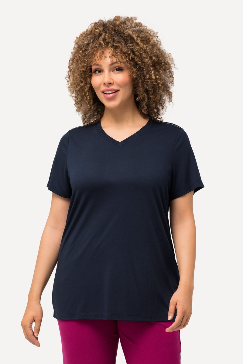 Donna con capelli ricci che indossa una maglietta blu navy a V con maniche corte e pantaloni magenta, in piedi davanti a uno sfondo chiaro e uniforme.