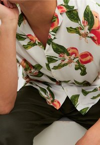 Camisa blanca de manga corta con un vibrante estampado de hojas en melocotón y verde. Presenta un cuello con solapa y botones, hecha de un tejido ligero.