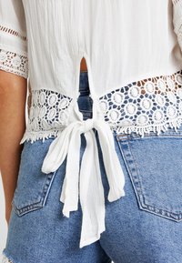 Blusa bianca con merletto in vita, dotata di un'apertura a chiave sul retro e dettagli con lacci. Abbinata a pantaloni corti in denim blu.