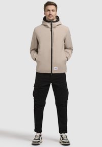 Jachetă waterproof bej cu glugă și fermoar frontal, cu manșete elastice și un patch cu logo, asortată cu pantaloni cargo negri și adidași.