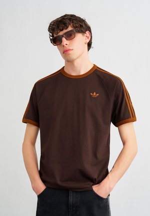Młody mężczyzna o kręconych włosach, ubrany w brązowy t-shirt Adidas, ciemne okulary przeciwsłoneczne i czarne spodnie, stoi z rękami w kieszeniach.