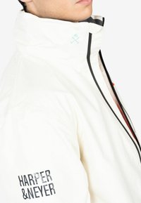Chaqueta impermeable blanca con un cuello alto, detalles de cremallera negra y un logo gráfico en la manga izquierda. Tejido ligero con un diseño minimalista.