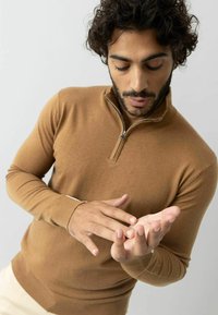 Hnedý sveter na zips z hladkého materiálu, s dlhými rukávmi a rebrovanými manžetami. Model skúma svoju ruku na neutrálnom pozadí.