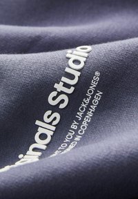 Tmavomodrá látka s bielym textom „Originals Studio“, s hladkou textúrou a jemným leskom. Text je výrazne zobrazený na povrchu.