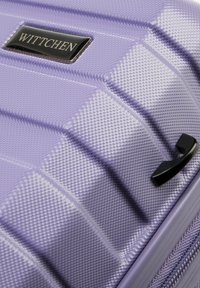 Maleta rígida morada con acabado texturizado, que presenta una placa de logo negra y un asa negra, destacando un diseño moderno.