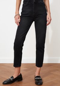 Jeans negros de talle alto con pierna cónica y bolsillos frontales, combinados con mocasines negros que tienen un detalle de cadena plateada, sobre un suelo de madera.