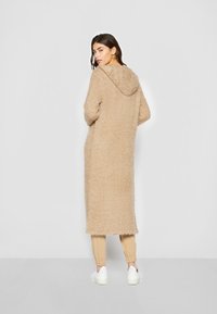 Beige, fluffy lang frakk med hette, med myk tekstur, ribbede kanter og kne-lengde design, båret over avslappede beige bukser og hvite sneakers.