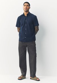 Camicia a maniche corte blu navy con bottoni, pantaloni grigi e sandali verde oliva. Caratterizzata da una vestibilità rilassata e una trama del tessuto liscia.