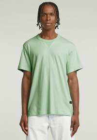 T-shirt di cotone verde chiaro con maniche corte, scollo rotondo e un sottile dettaglio a V. Presenta un piccolo tag con il marchio all'orlo.