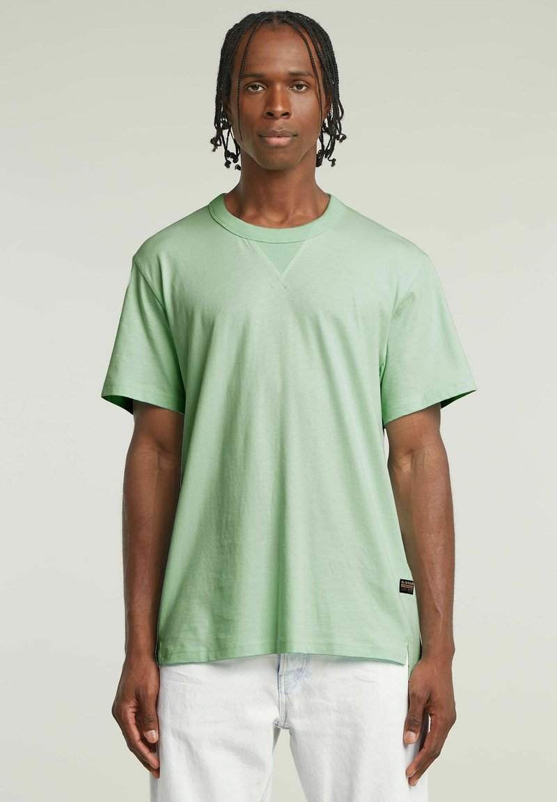 T-shirt di cotone verde chiaro con maniche corte, scollo rotondo e un sottile dettaglio a V. Presenta un piccolo tag con il marchio all'orlo.