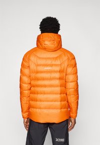 Casaco puffer laranja com padrão de grelha, capuz e logótipo nas costas. O material parece acolchoado e texturizado para isolamento.