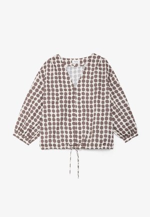 Vrouw met kort bruin haar draagt zilveren oorringen en een crèmekleurige blouse met grote bruine stippen en een V-hals. Neutrale achtergrond.
