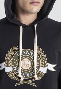 Sudadera negra con cordones blancos, que presenta un logo bordado en dorado y verde con acentos de laurel y el año de fundación, 2017.