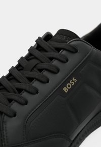 Gros plan d'une basket en cuir noir avec des lacets noirs et le logo doré "BOSS" sur le côté, montrant un panneau en tissu texturé et les détails de la semelle.
