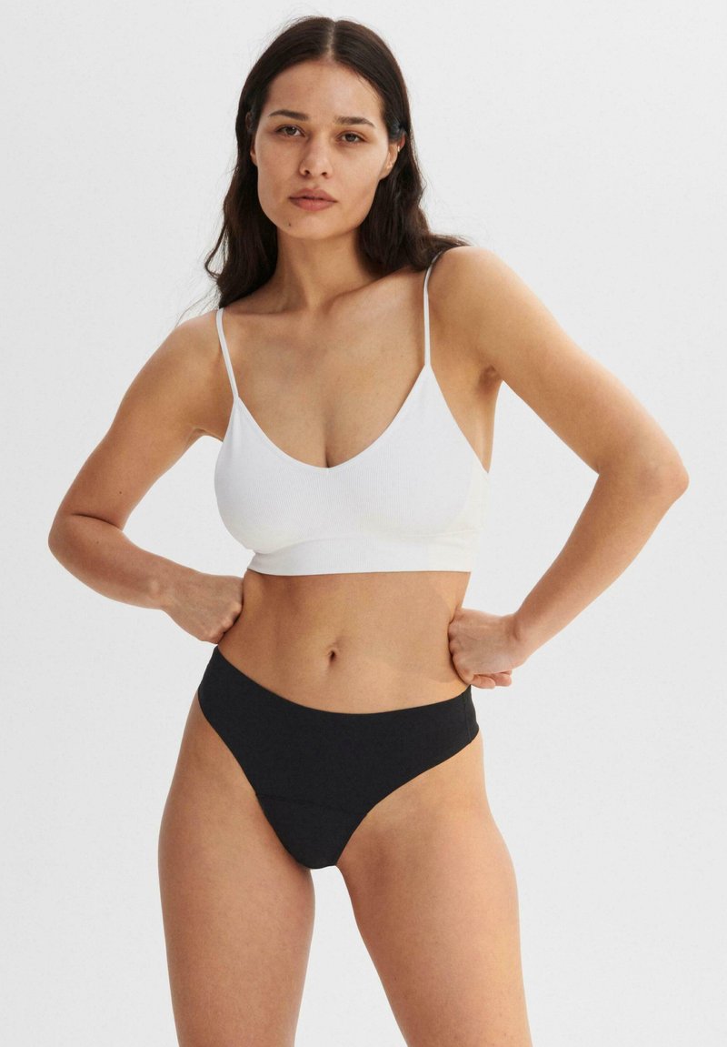 Weißer gerippter Crop-Top mit dünnen Trägern und tiefem V-Ausschnitt, kombiniert mit schwarzen hochgeschnittenen Bikinihosen aus glattem Material und minimalen Nähten.