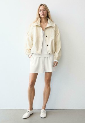 Donna in piedi contro un muro bianco che indossa una giacca beige, un top lavorato a maglia bianco, pantaloncini bianchi e scarpe bianche con lacci, con le mani nelle tasche.