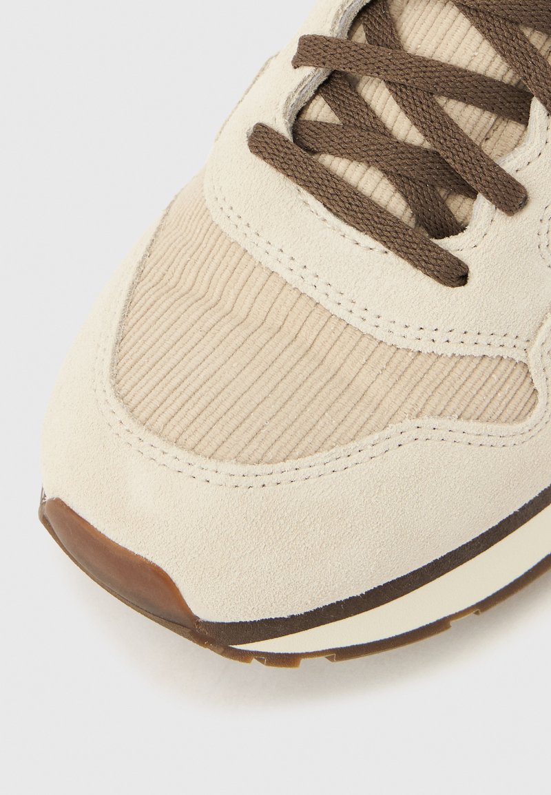 Baskets en daim beige avec accents en velours côtelé, bout rond, lacets marron et semelle intermédiaire blanche. Surface texturée et détails de couture visibles.