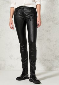 Osoba ubrana w błyszczące czarne dopasowane spodnie, czarne lakierowane botki i białą koszulę z podwiniętymi rękawami, stojąca na jasnej podłodze.