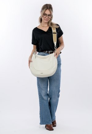 Weiße Leder-Schultertasche mit geschwungener Form, verstellbarem Tragegurt und silbernem Hardware, gehalten von einer Person in einem schwarzen Oberteil und blauen Jeans.