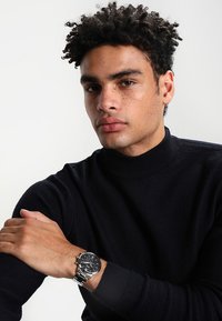Pull en maille noire avec un col haut. Montre argentée au poignet avec un cadran noir. Le modèle a des cheveux bouclés et une expression neutre.