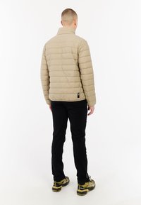 Man die met zijn rug naar de camera staat, gekleed in een beige gewatteerde jas, zwarte broek en gele wandelschoenen voor een witte achtergrond.