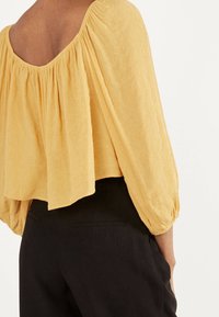 Żółta bluzka o fakturze z szerokim, zaokrąglonym dekoltem i bufkami, zestawiona z czarnymi, dopasowanymi spodniami. Krawędzie są lekko nierówne.