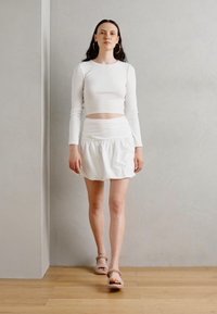 Balta ilgomis rankovėmis trumpa palaidinė derinama su balta raukšlėta mini sijonu, pasižyminčiu lygiu paviršiumi. Modelis dėvi minimalistinius sandalus.
