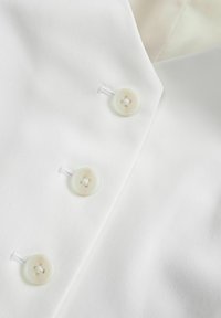 Zbliżenie na białą odzież, na którym widać trzy przezroczyste białe guziki oraz część sztywnego kołnierzyka z subtelną teksturą.