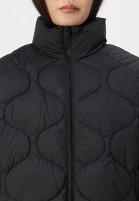 Svart quiltad pufferjacka med hög krage, utrustad med dragkedja fram och rundad, vågformad sömnadsdesign. Mjuk textur.