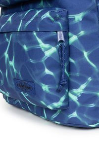 Sac à dos en tissu bleu avec un motif vert semblable à de l'eau, présentant une poche frontale et un patch de logo. Comprend un détail en cordon sur la fermeture éclair.