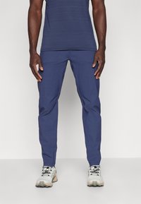 Marineblå bukser med slim fit, der har en glat overflade, to frontlommer og et tapered ben-design. Kombineret med grå og beige sko.
