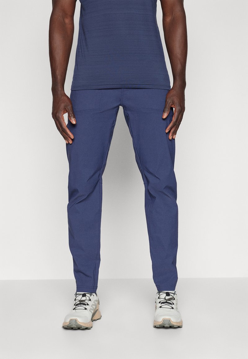 Marineblå bukser med slim fit, der har en glat overflade, to frontlommer og et tapered ben-design. Kombineret med grå og beige sko.
