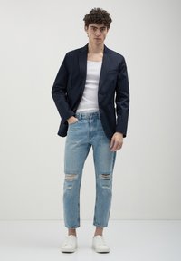 Blazer blu navy, canotta bianca, jeans leggeri azzurri strappati e sneakers bianche. Il blazer ha un bottone singolo e una tasca frontale.