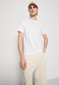 Hvit t-skjorte med korte ermer og rund hals, med liten brodert logo på brystet, kombinert med beige vide bukser og en kremfarget caps.