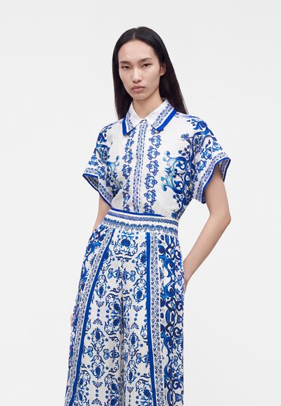 Femme aux longs cheveux noirs portant un chemisier à manches courtes blanc et bleu à motif floral et un pantalon large assorti sur un fond uni.