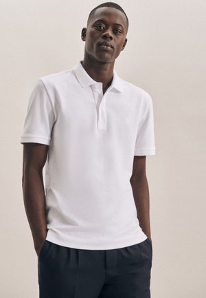 Hvid kortærmet polo-shirt med krave og tre-knaps lukning, lavet af glat stof. Båret over mørke bukser, hænderne i lommerne.